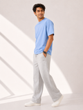 A man wearing a light blue crewneck t-shirt, light gray sweatpants, and white sneakers, standing in a brightly lit studio with soft diagonal shadows.