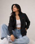 A woman sits cross-legged wearing a black zip-up bomber jacket with a small red embroidered logo on the chest, a white crop top, light-wash blue jeans, and white sneakers against a plain light gray studio background.
