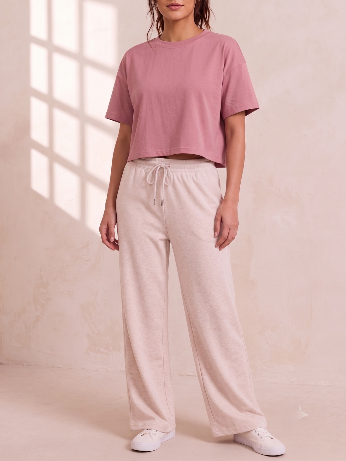A person wearing a dusty pink cropped short-sleeve crewneck t-shirt and light beige drawstring sweatpants, photographed in soft natural light with window shadows.