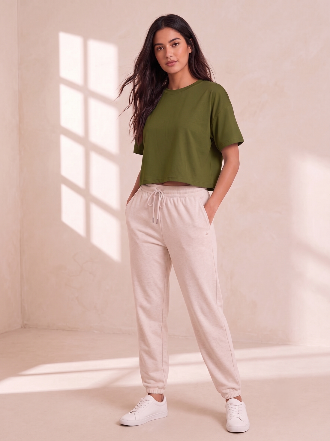 A woman wearing an olive green short-sleeve crop top and light beige drawstring sweatpants, shown from the neck down against a softly lit neutral background.