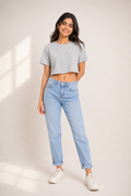A woman modeling a light gray cropped t-shirt and light blue straight-leg jeans with white sneakers, standing against a warm beige studio background with soft window shadow projections.