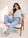 A woman sitting on a wooden armchair wearing a light gray short-sleeve crop top, light blue high-waisted jeans, and white sneakers in a brightly lit studio setting.