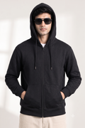 A man wearing a black full-zip hoodie with a drawstring hood, black sunglasses, and light beige pants, standing against a light gray background.