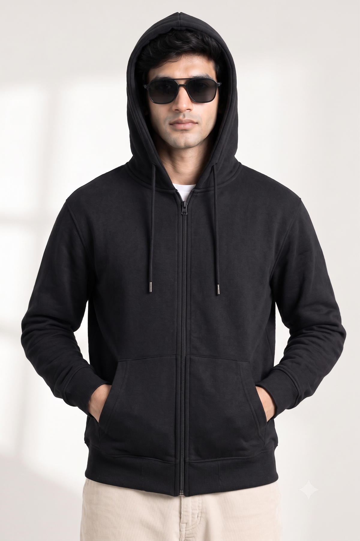 A man wearing a black full-zip hoodie with a drawstring hood, black sunglasses, and light beige pants, standing against a light gray background.