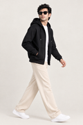 A man wearing a black zip-up hoodie, white t-shirt, beige trousers, and white sneakers walking against a plain light beige studio background.