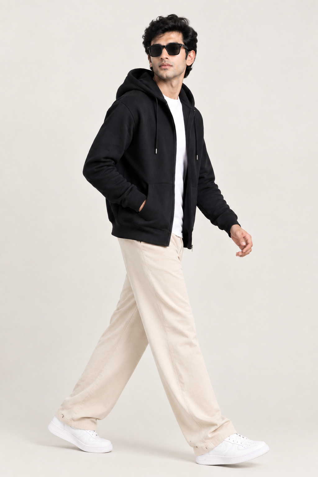 A man wearing a black zip-up hoodie, white t-shirt, beige trousers, and white sneakers walking against a plain light beige studio background.