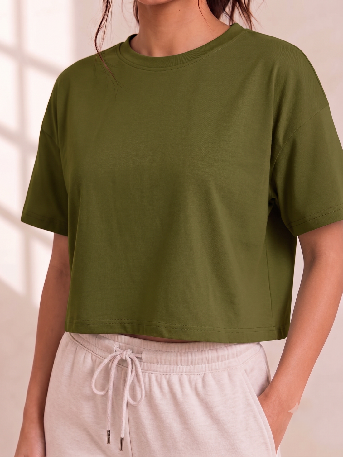 A woman wearing an olive green short-sleeve crop top and light beige drawstring sweatpants, shown from the neck down against a softly lit neutral background.