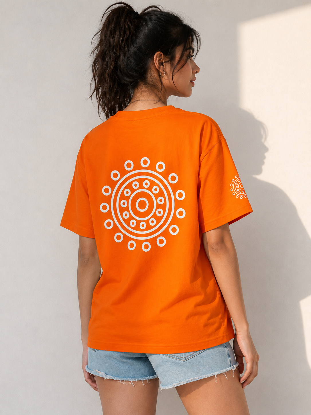A woman shown from the back wearing a bright orange t-shirt with a large white circular mandala-style graphic on the back and a smaller matching graphic on the left sleeve, paired with light blue denim shorts against a neutral wall.