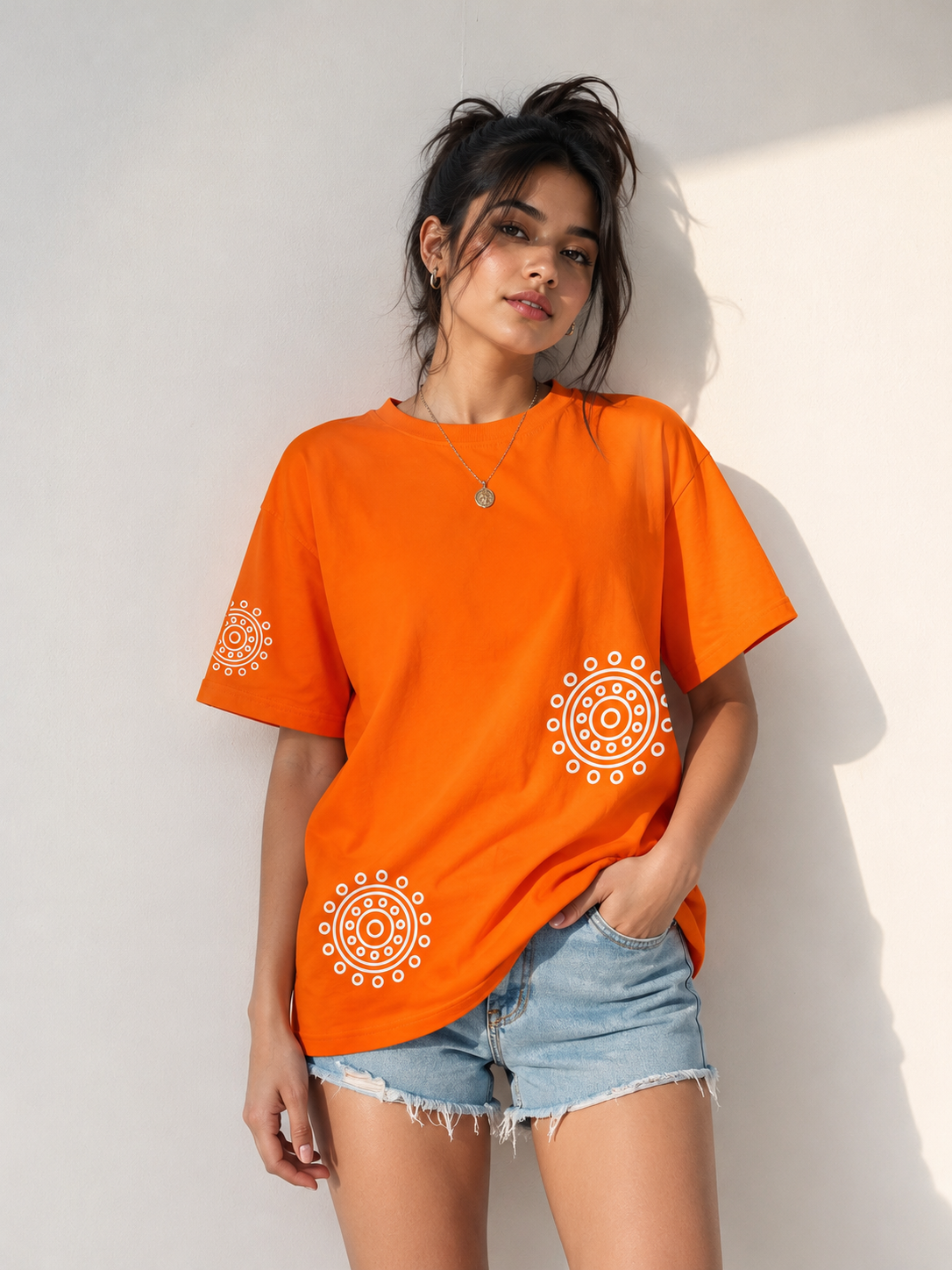 A woman wearing an oversized bright orange t-shirt with white circular mandala prints on the front and sleeves, paired with light-wash denim shorts, posing against a white wall with soft shadows.