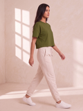 A woman walking in profile wearing a short-sleeve olive green crop top, cream drawstring jogger pants, and white sneakers in a softly lit room with warm sunlight and window shadows.