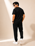 A man wearing a black short-sleeve t-shirt, black jogger pants, and white sneakers, photographed from the back in a studio setting with warm beige walls and floor shadows.