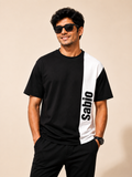 A man wearing a black and white color-block t-shirt with 'Sabio' printed vertically in black on the white panel, paired with black pants, against a solid beige background.
