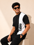 A man wearing a black and white color-blocked t-shirt with the brand name 'sabio' printed vertically in black on the white panel, paired with black pants, sitting against a beige background.