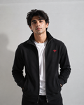 A man wearing a black zip-up fleece jacket with a small red embroidered logo on the left chest, layered over a white t-shirt, standing against a light gray studio background.