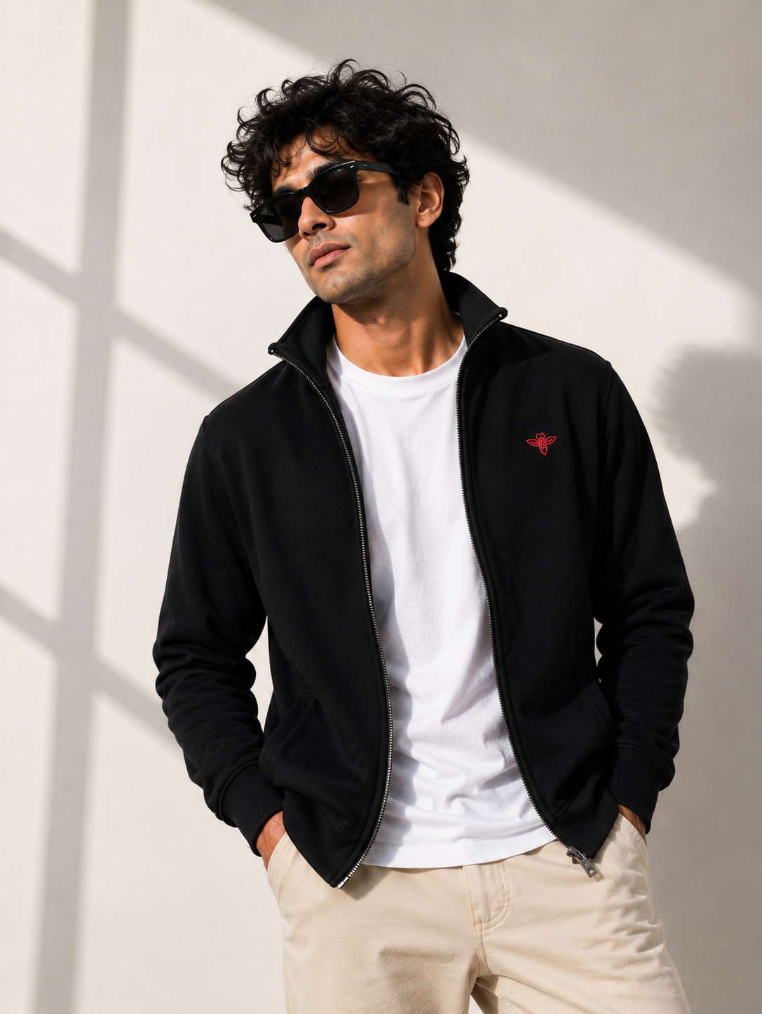 A man wearing a black zip-up fleece jacket with a small red embroidered logo on the chest, layered over a white t-shirt and beige pants, standing in front of a wall with window shadows.