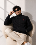 A man wearing a black zip-up jacket with a small red embroidered logo on the chest, beige trousers, and white sneakers, sitting in a beige armchair and wearing black sunglasses.