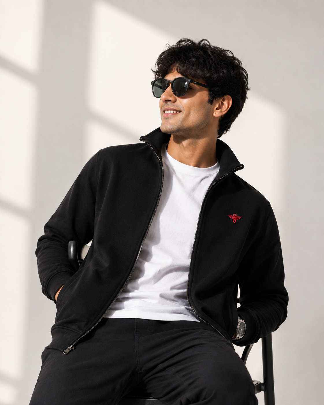 A man wearing a black zip-up fleece jacket with a small red embroidered logo on the chest, a white t-shirt, and black pants, sitting against a white wall with soft window shadows.