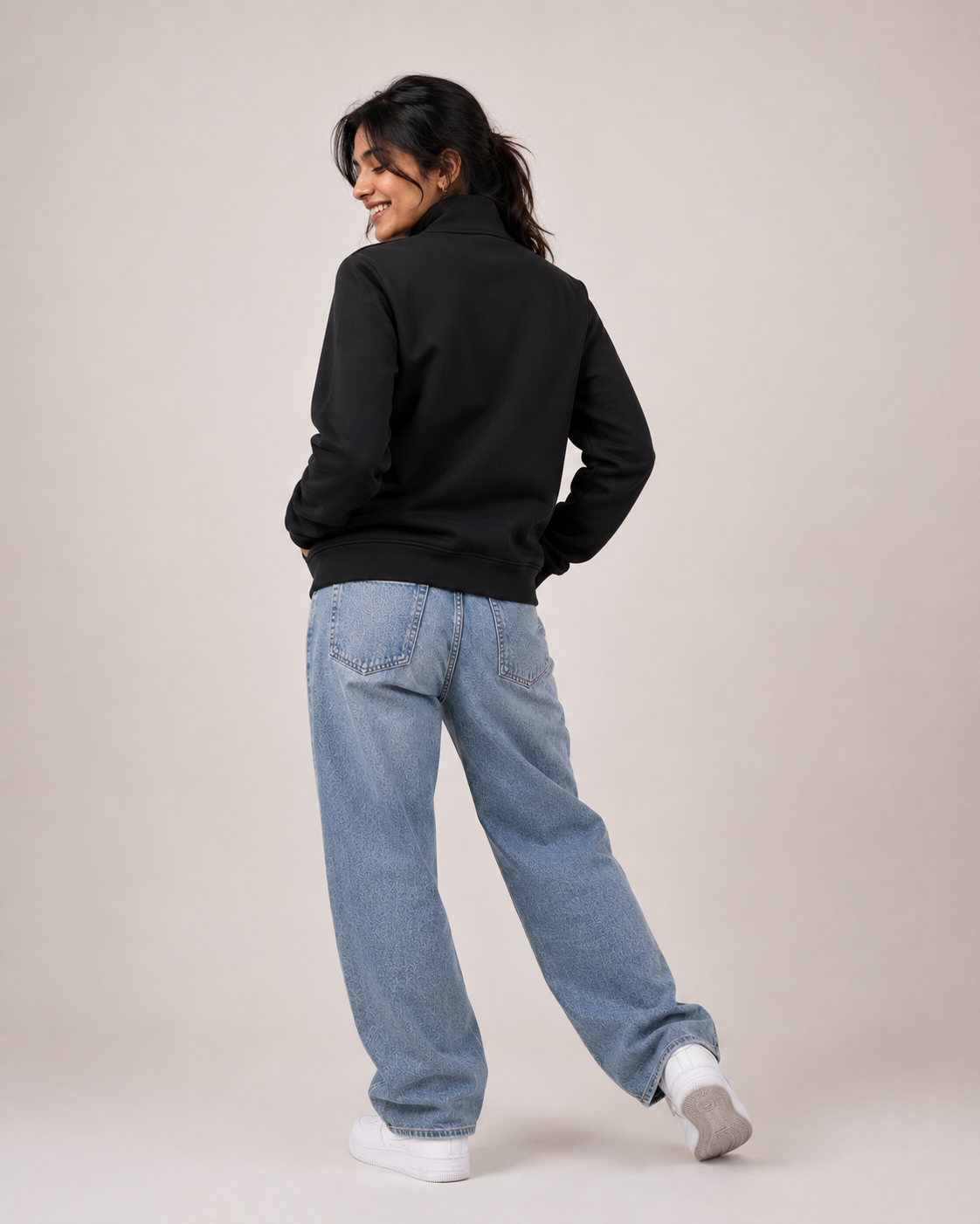 A woman wearing a black zip-up jacket, light blue wide-leg jeans, and white socks, photographed from behind against a plain beige studio background.