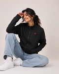 A woman sitting on the floor wearing a black zip-up hoodie with a small red embroidered logo on the left chest, paired with light blue jeans and white sneakers, against a plain light beige background.
