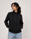A woman wearing a black zip-up hoodie with a small red embroidered logo on the chest, paired with light blue jeans, photographed against a plain light gray studio background.