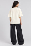 A woman shown from the back wearing an oversized cream-colored t-shirt and wide-leg black pants, standing against a light gray studio background.