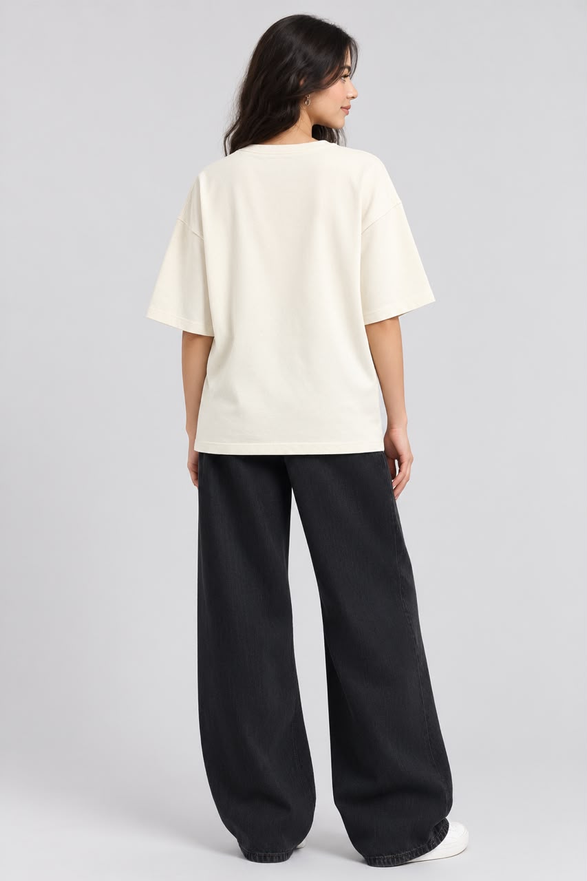A woman shown from the back wearing an oversized cream-colored t-shirt and wide-leg black pants, standing against a light gray studio background.
