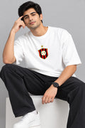 A man wearing a white crewneck t-shirt with a red and black striped shield crest on the chest, paired with black jeans and white sneakers, sitting on a white block against a gray background.