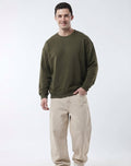 A man wearing an olive green crewneck sweatshirt and beige pants, standing against a plain white studio background.
