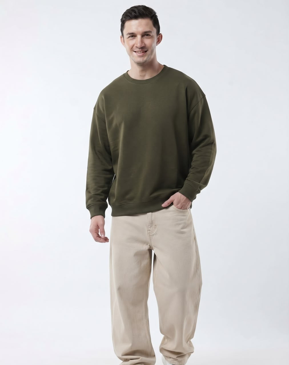 A man wearing an olive green crewneck sweatshirt and beige pants, standing against a plain white studio background.