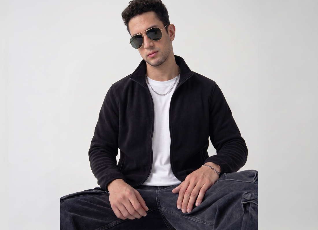 A man wearing a black zip-up fleece jacket over a white t-shirt and dark jeans, sitting against a light gray background.