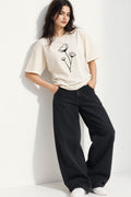 A woman models a cream-colored short-sleeve t-shirt with a black line-art flower graphic on the front, paired with black wide-leg jeans and white sneakers, leaning against a white wall.