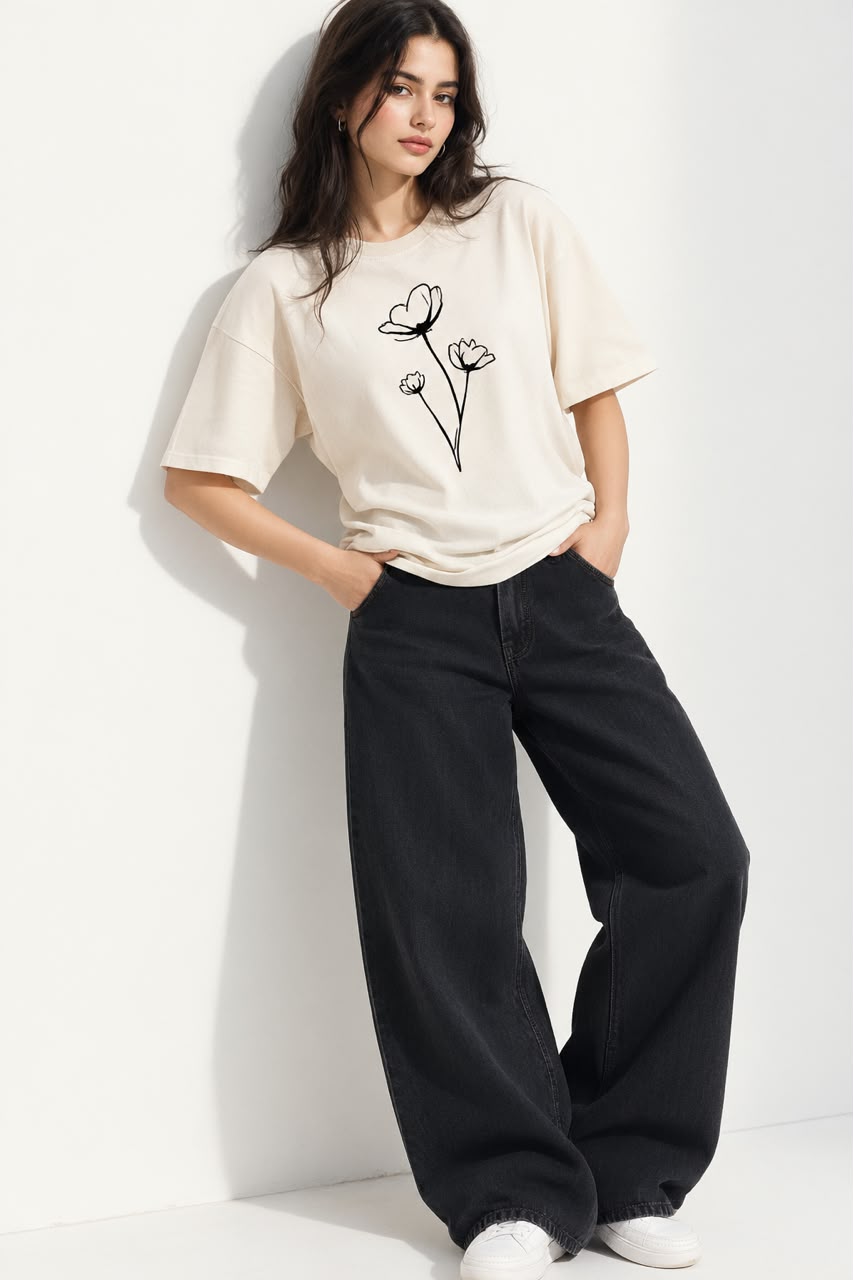 A woman models a cream-colored short-sleeve t-shirt with a black line-art flower graphic on the front, paired with black wide-leg jeans and white sneakers, leaning against a white wall.