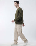 A man in profile wearing an olive green crewneck sweatshirt, beige relaxed-fit trousers, and white sneakers, walking against a plain light gray studio background.