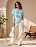 A woman stands in a sunlit indoor setting wearing a light blue graphic T-shirt with the words 'KEEP TIME' in beige and gray, paired with wide-leg cream pants, white sneakers, and a cream shoulder bag.