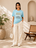 A woman stands in a sunlit indoor room wearing a light blue graphic T-shirt with the words 'KEEP LIFE SIMPLE' in muted gold and gray, paired with off-white cargo pants and white sneakers. She holds a small light-colored handbag.