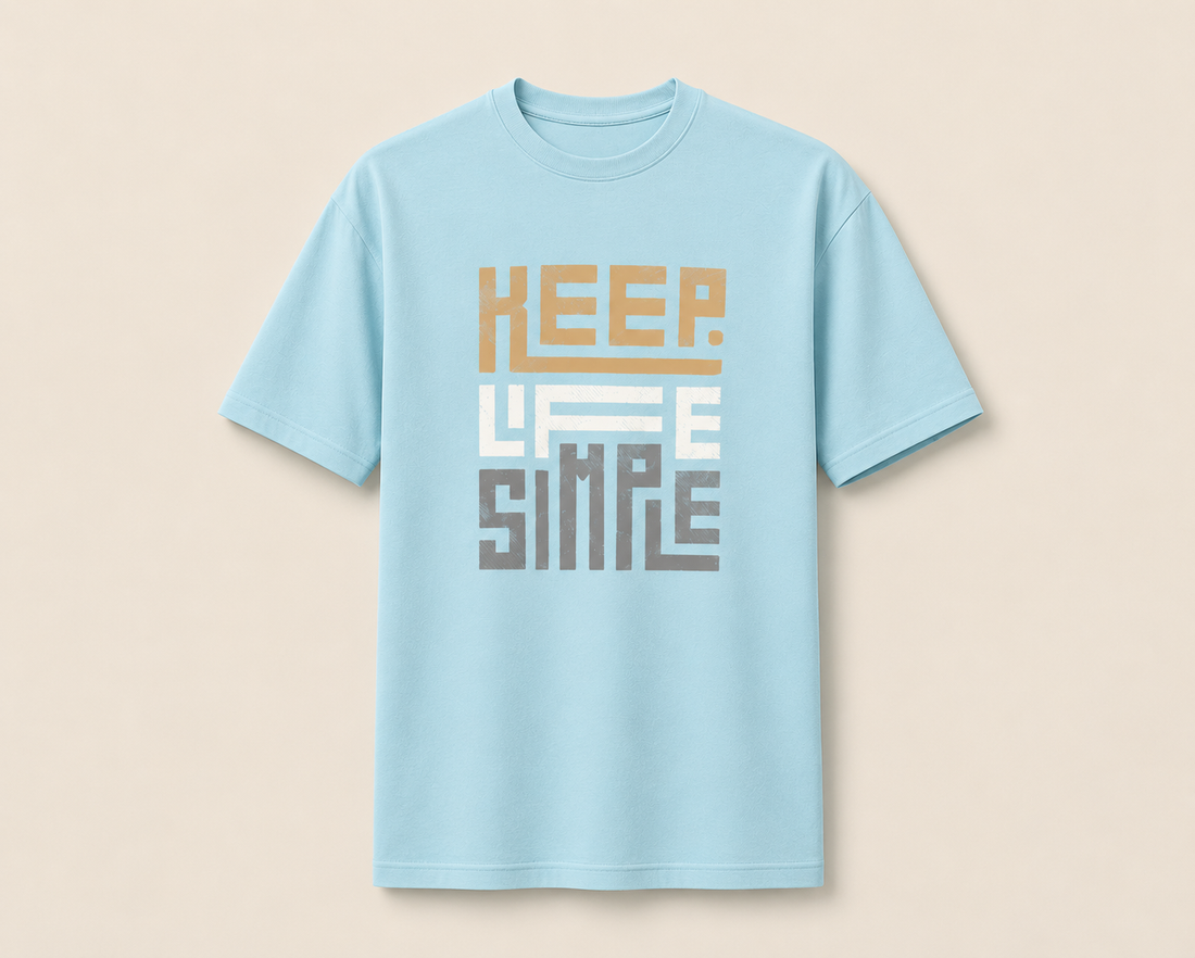 A light blue short-sleeve crewneck t-shirt with a retro-style graphic on the chest reading 'KEEP LIFE SIMPLE' in stacked blocks of tan, white, and gray, displayed against a solid beige background.