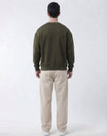 A man viewed from behind wearing an olive green crewneck sweatshirt and beige trousers, standing against a light gray studio background.