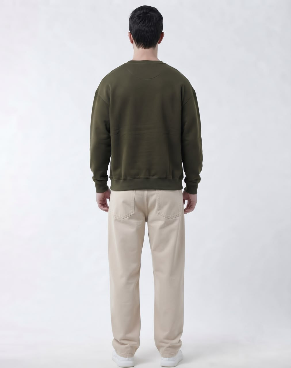 A man viewed from behind wearing an olive green crewneck sweatshirt and beige trousers, standing against a light gray studio background.