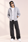 A man modeling a light gray zip-up hoodie over a white t-shirt, paired with black wide-leg jeans and white sneakers, standing against a warm beige background.
