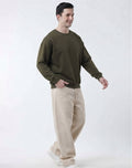 A man wearing an olive green crewneck sweatshirt and beige wide-leg trousers, walking against a plain white studio background.