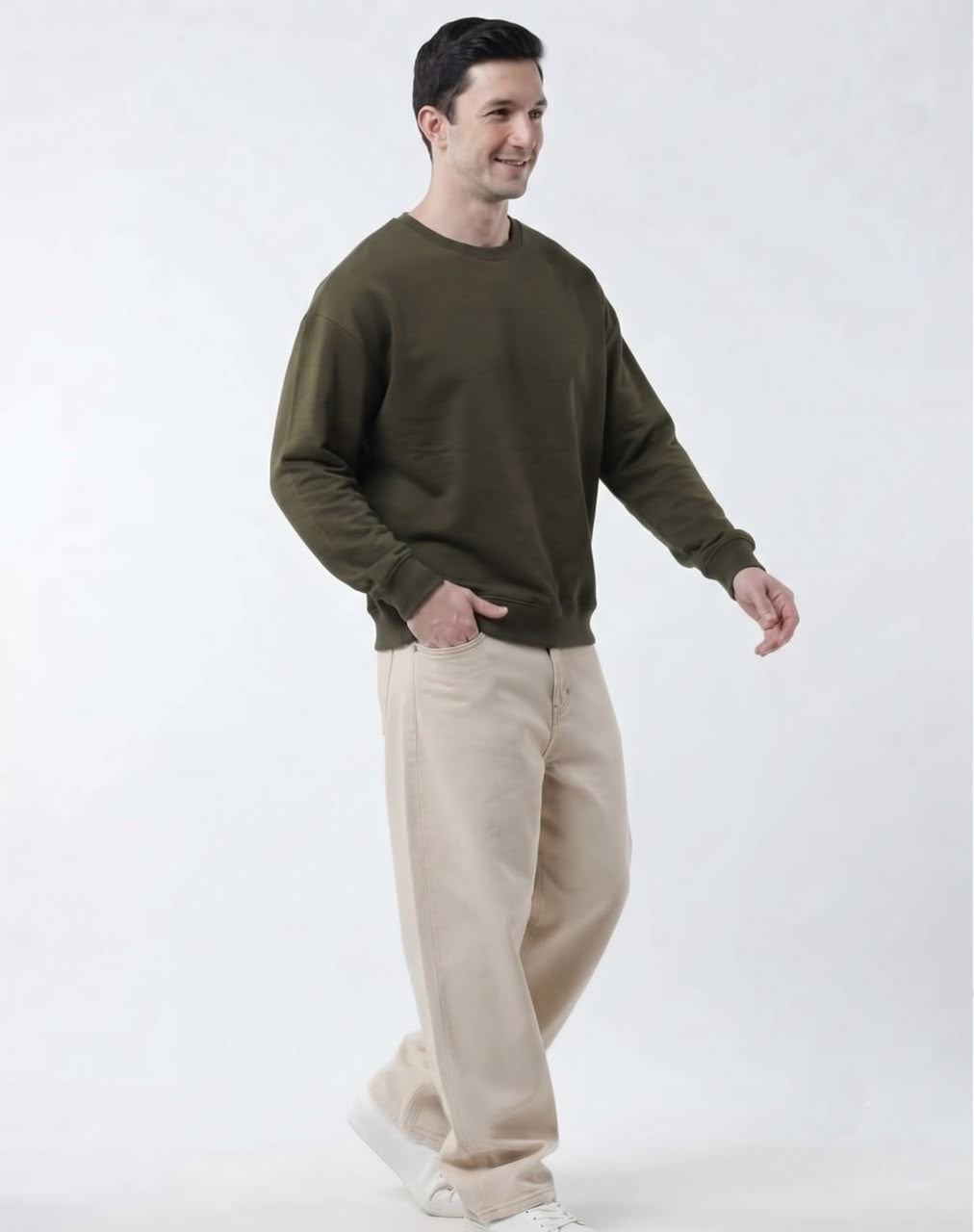 A man wearing an olive green crewneck sweatshirt and beige wide-leg trousers, walking against a plain white studio background.