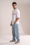 Full-body studio shot of a smiling Indian male model wearing The Sabio's oversized white RELOVEUTION graphic t-shirt, baggy light blue denim jeans, and casual sneakers, facing forward.