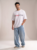 Side profile shot of a male model wearing The Sabio oversized white RELOVEUTION t-shirt and baggy light blue denim jeans in a studio setting, showcasing the loose-fitting drop-shoulder silhouette.