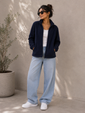 A woman wearing a navy blue zip-up fleece jacket, light-wash wide-leg jeans, a white tank top, and white sneakers, standing against a textured beige wall with a potted plant on the left.