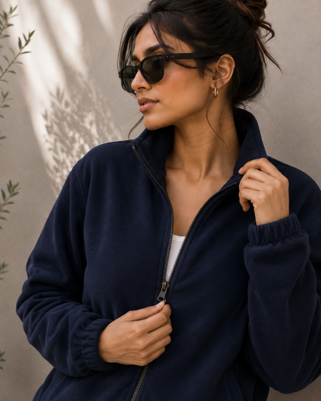A woman wearing a navy blue fleece zip-up jacket, black sunglasses, and gold hoop earrings, posing against a textured beige wall with leaf shadows.