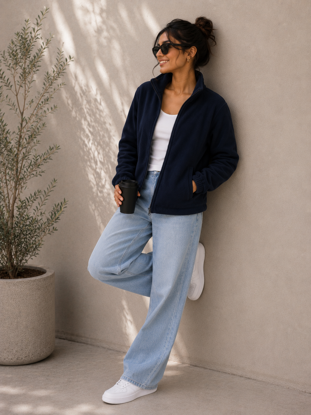 A woman wearing a navy blue fleece zip-up jacket, black sunglasses, and gold hoop earrings, posing against a textured beige wall with leaf shadows.